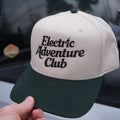 Cap with 'Electric Adventure Club' text held by a hand, blurred background